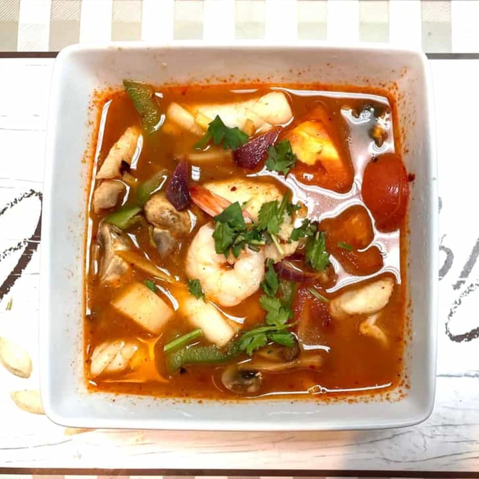 A white bowl of Thai Tom Yum soup with shrimp, mushrooms, tomatoes, and herbs.