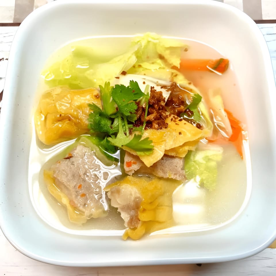 Thai clear soup with wontons, meatballs, cabbage, carrots, and cilantro in a white bowl.